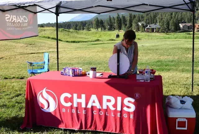A woman at a red table with the Charis Bible College logo, outdoors with a mountain backdrop.