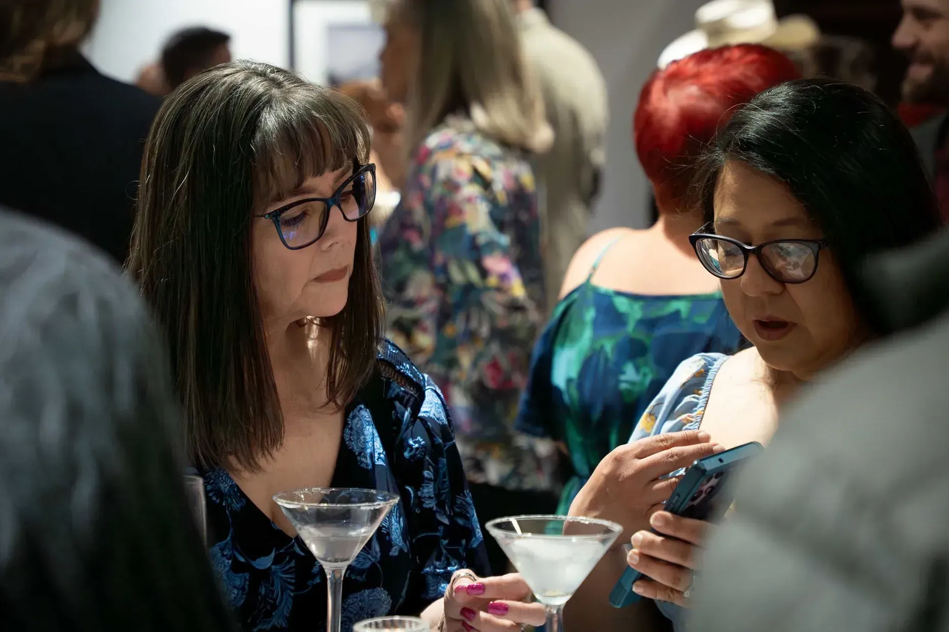 Two women in glasses at a gathering, one looks on at the other using a phone. Drinks are visible.