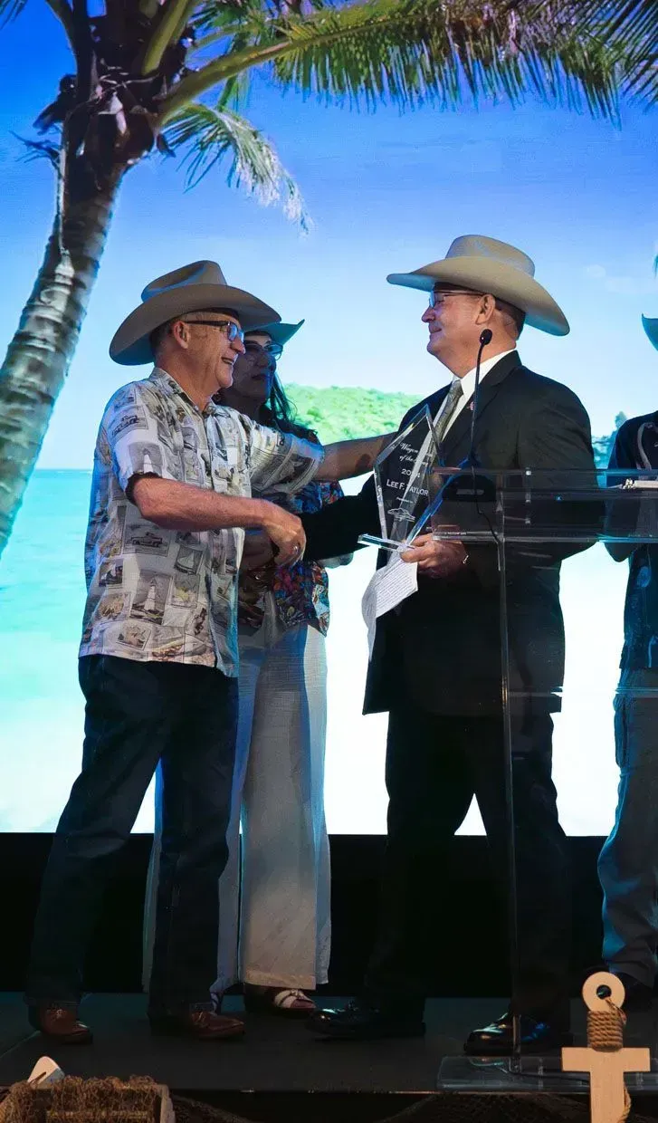 Two men in cowboy hats shake hands on a stage with a tropical backdrop.