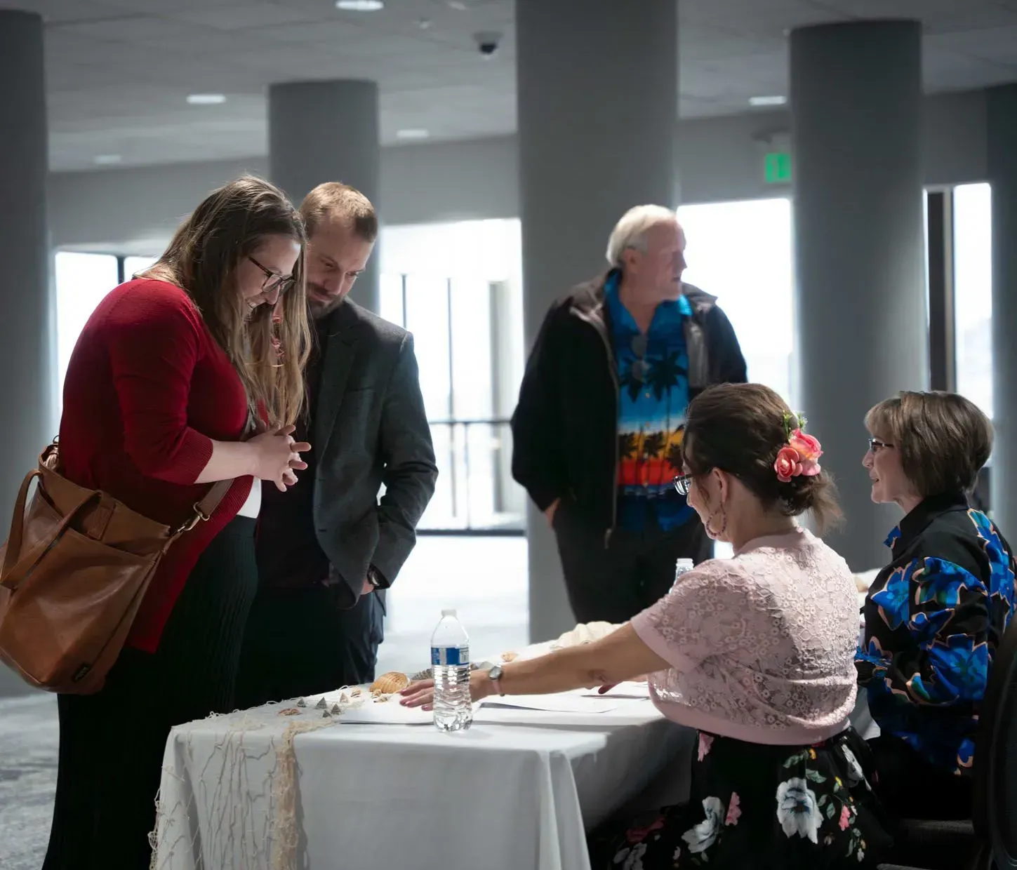 People gather at a table; a woman reads a hand. Others watch, in an event space with large windows and gray columns.