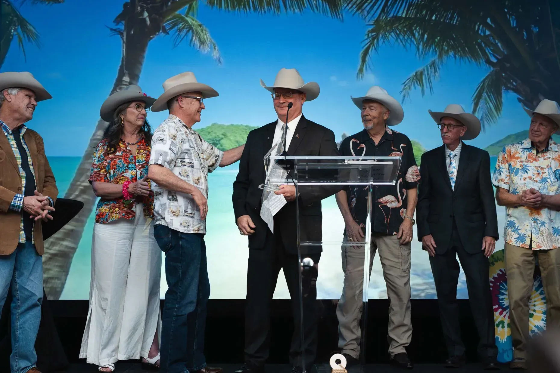 Group of people in cowboy hats and Hawaiian shirts on stage; man at podium.