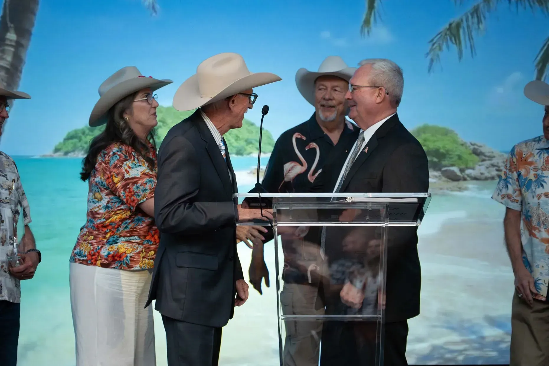 People in cowboy hats and suits shaking hands at a podium; tropical beach backdrop.