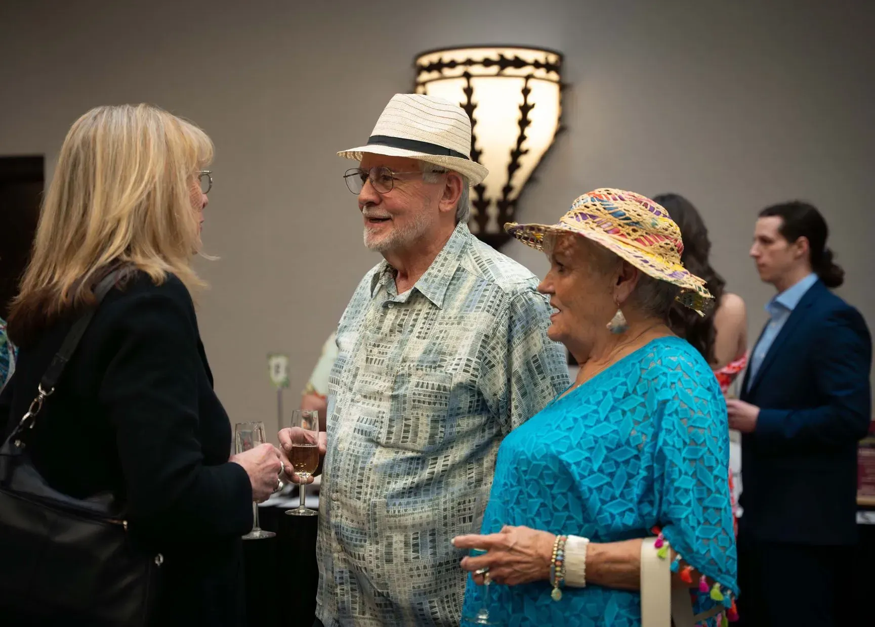 Three people in conversation at an event. Two wear hats, one in a blue shirt. Indoors, with a warm-lit fixture.