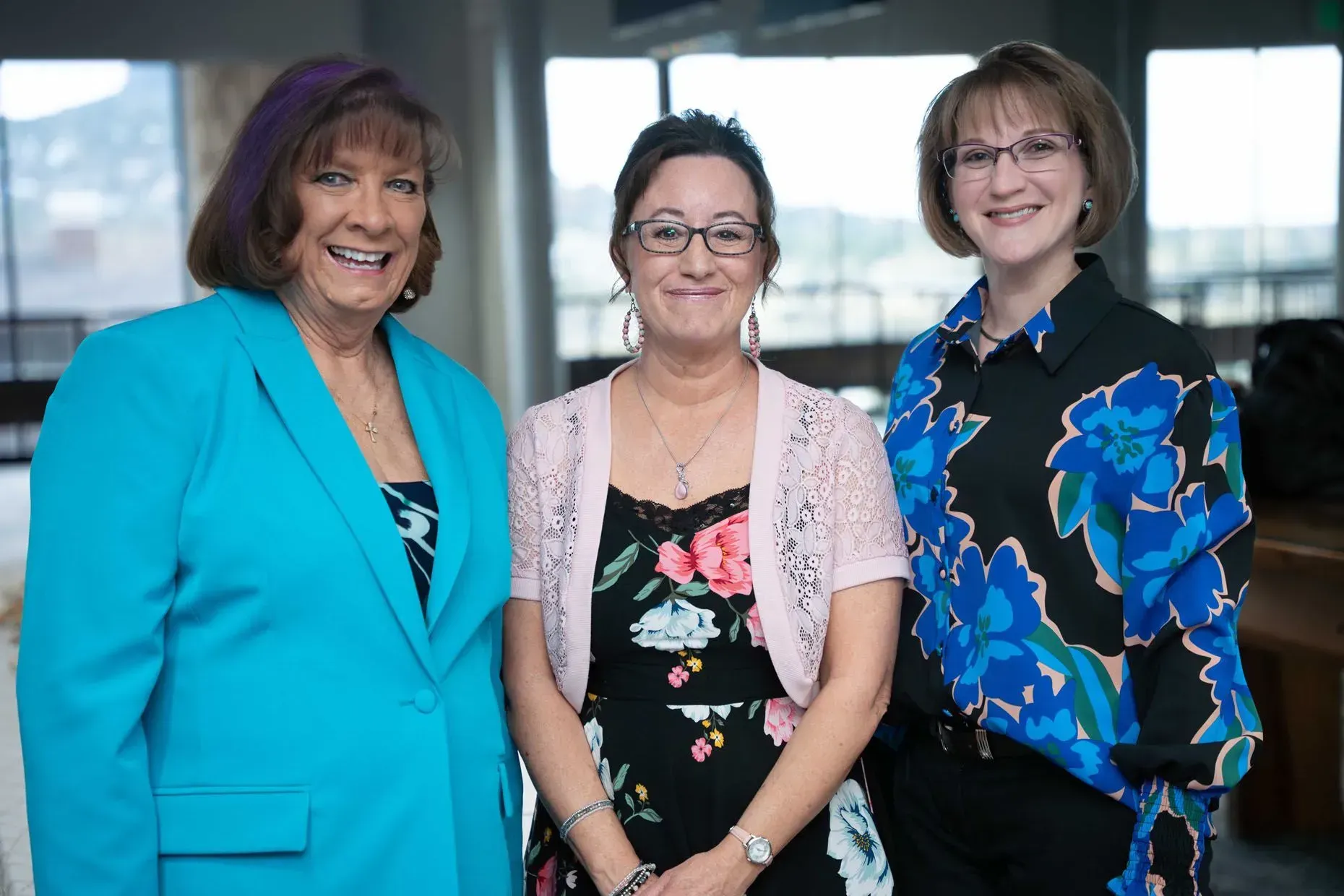 Three women smiling, posed indoors. Woman on left in teal blazer, center in floral dress and pink cardigan, right in floral shirt.