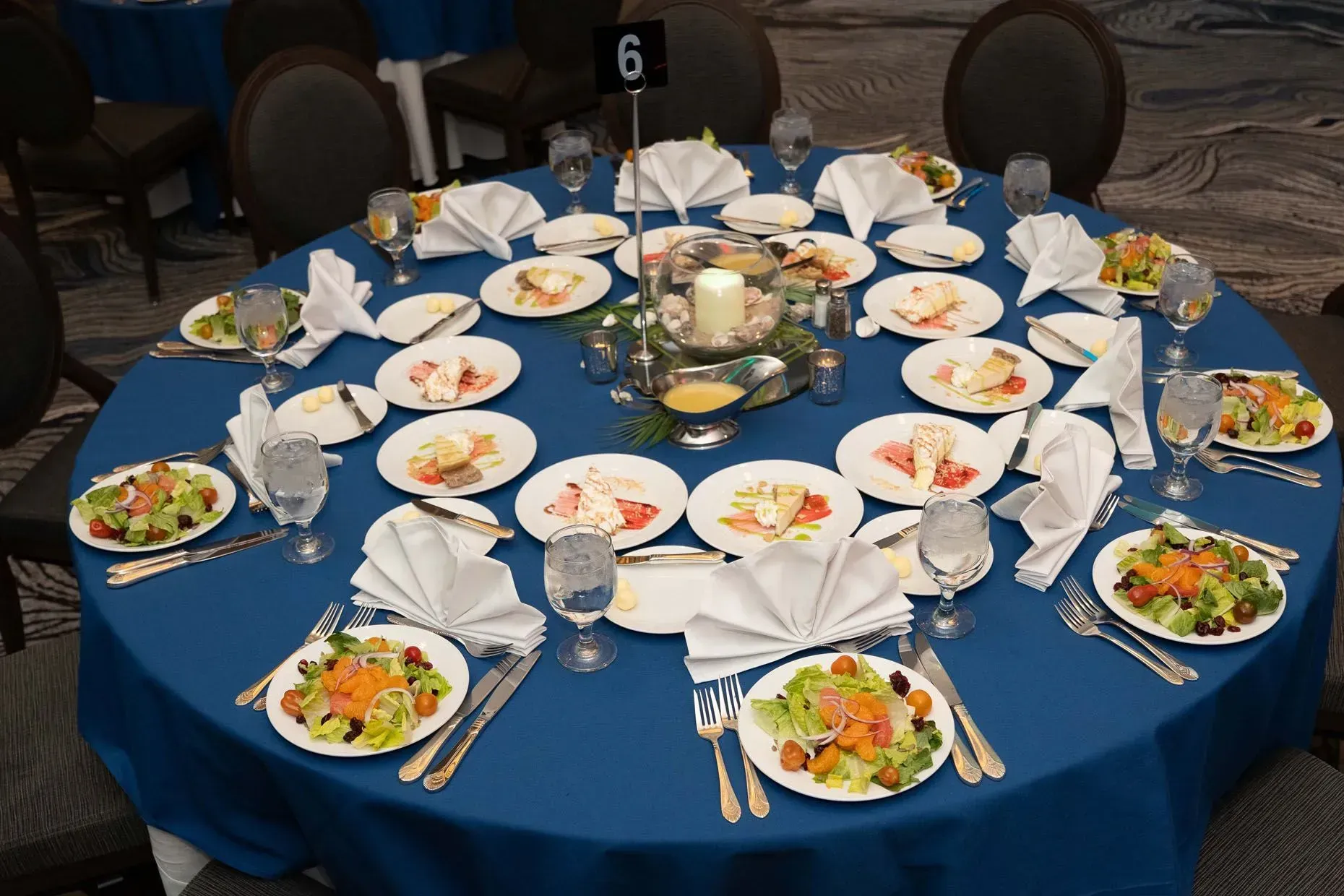 A round table set for a meal with plates, silverware, and salad, table number 6, blue tablecloth.