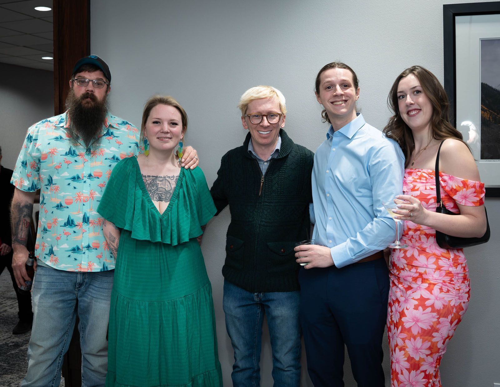 Five people smiling, posing indoors. Man with beard and floral shirt, woman in green dress, blonde man with arm around another, man in blue shirt, woman in floral dress.
