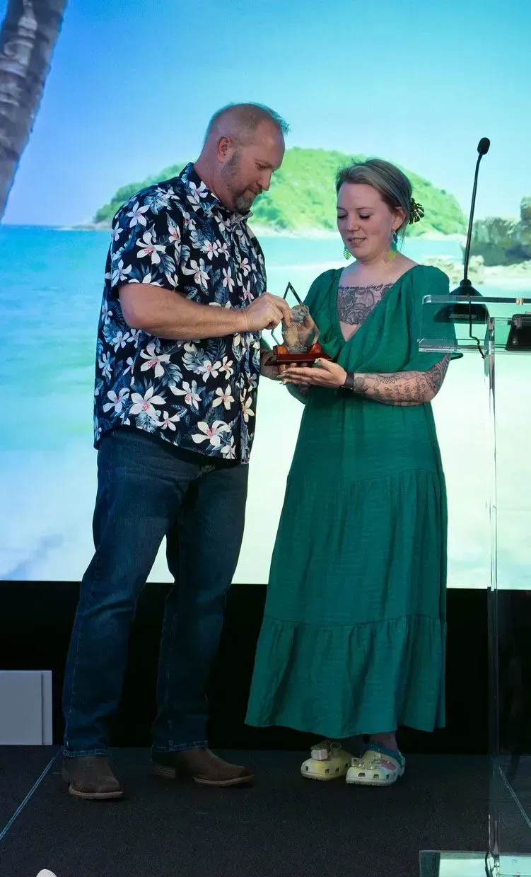 Man in Hawaiian shirt gives an award to woman in green dress, with beach background.