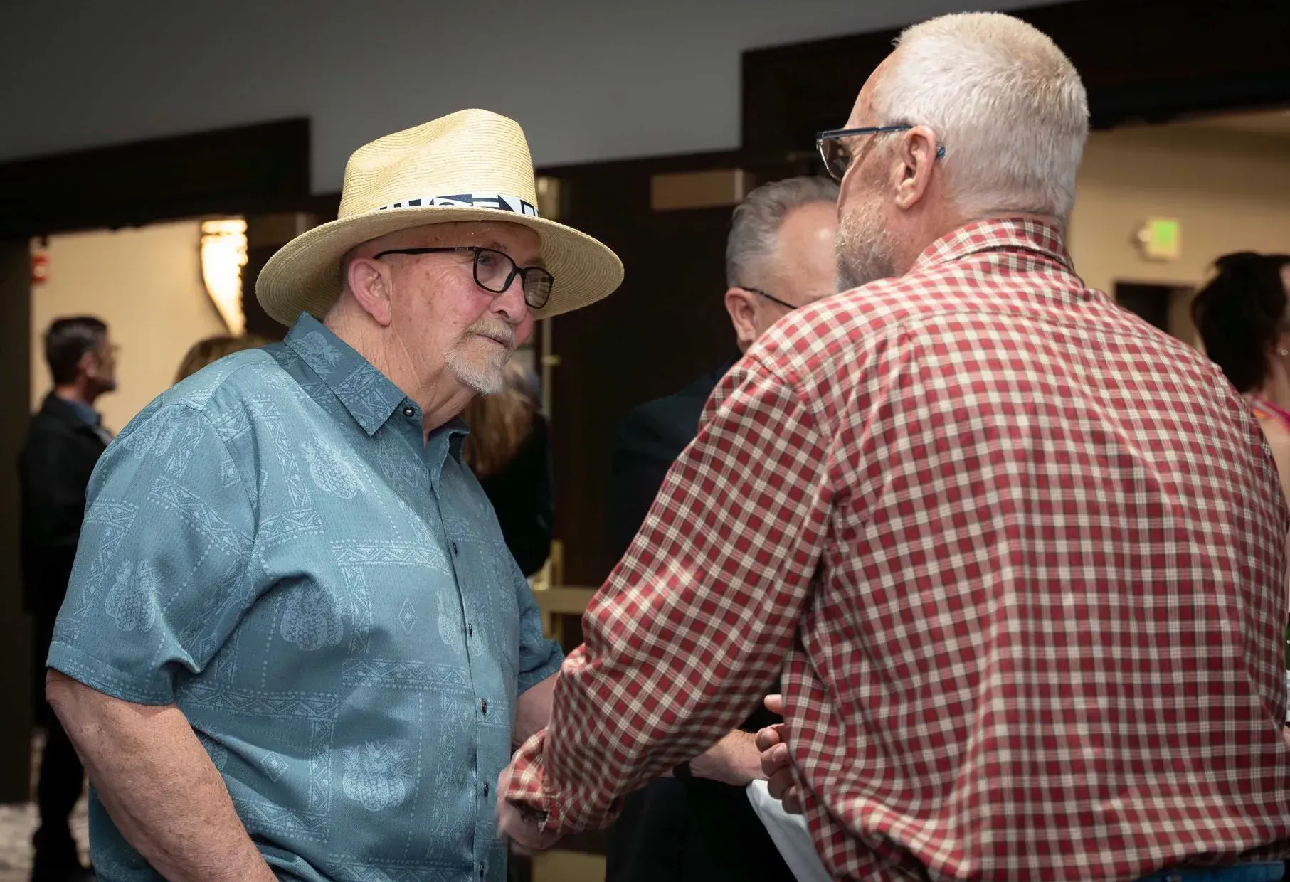 Two older men shaking hands in a room; one wears a straw hat and blue shirt, the other a plaid shirt.