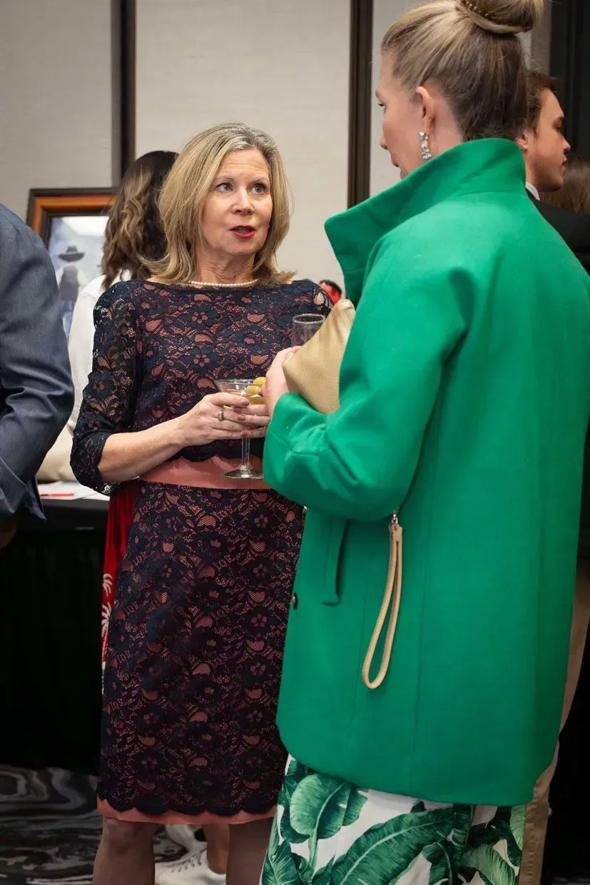 Woman in lace dress talking to woman in green coat, indoor setting.