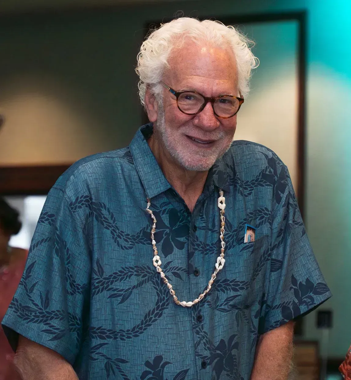 Older man with white curly hair, glasses, and blue floral shirt smiles, wearing shell necklace.