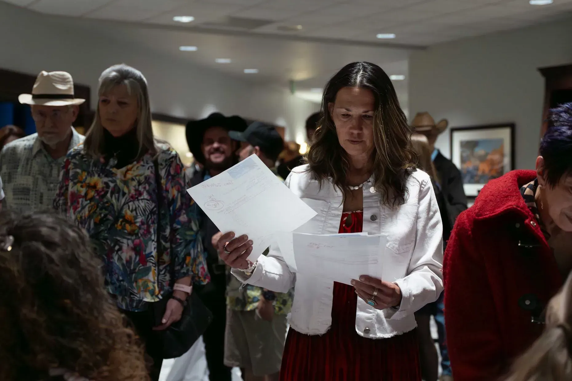 People in an event hall, one woman examines papers, others look on, some wearing hats.