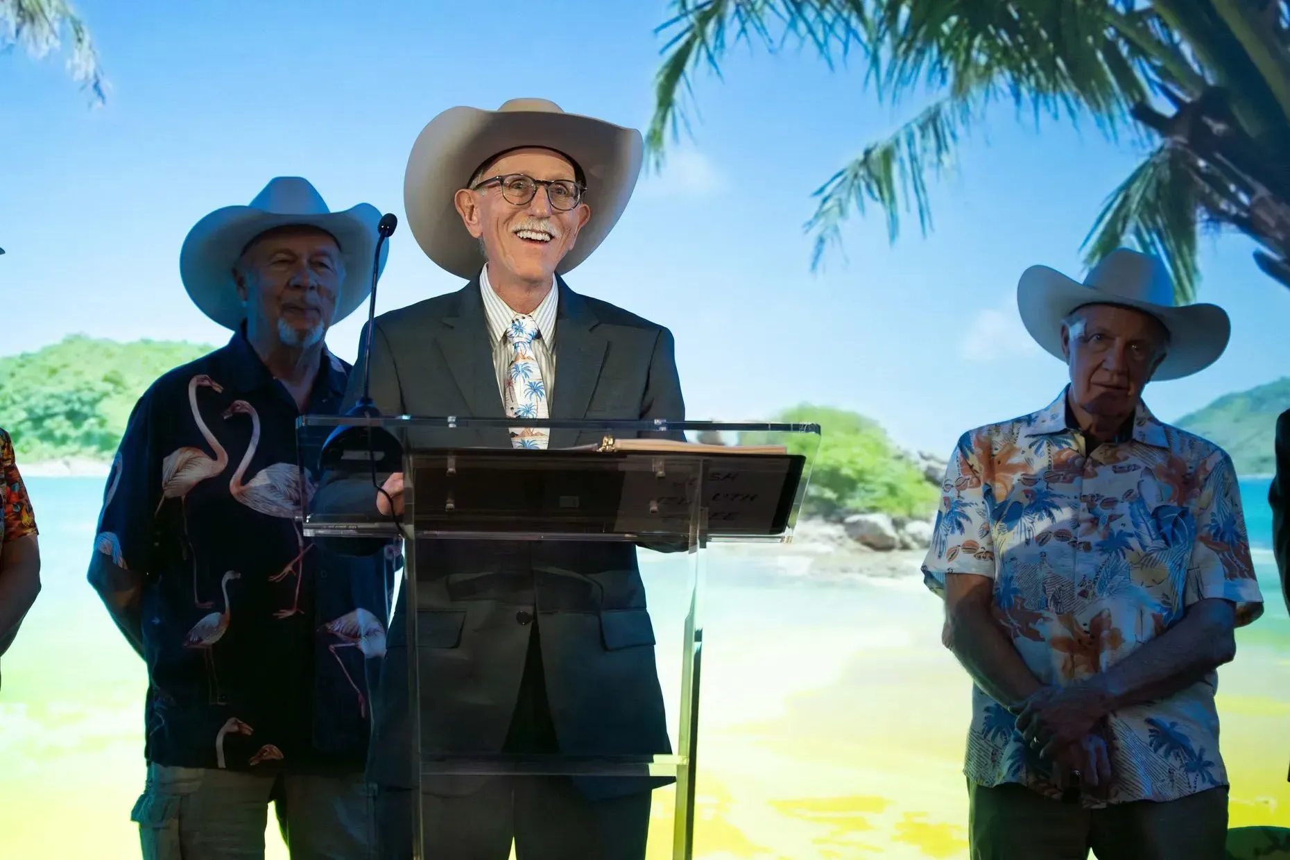 Three men in cowboy hats and Hawaiian shirts stand at a podium, smiling in front of a tropical backdrop.