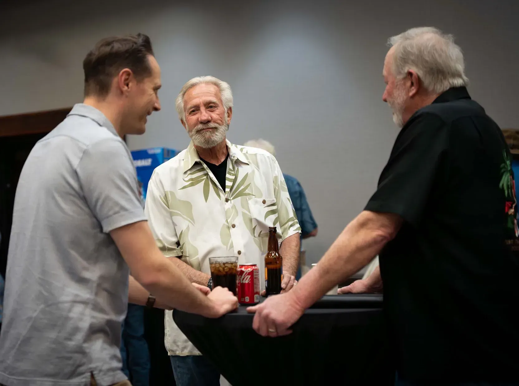 Three men converse near a table with drinks. One wears a floral shirt, another a grey shirt, and the last a black shirt.