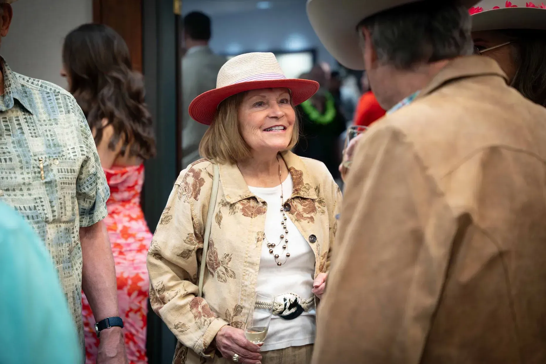 Woman in a beige jacket and red-trimmed hat chats at a gathering; other people are nearby.