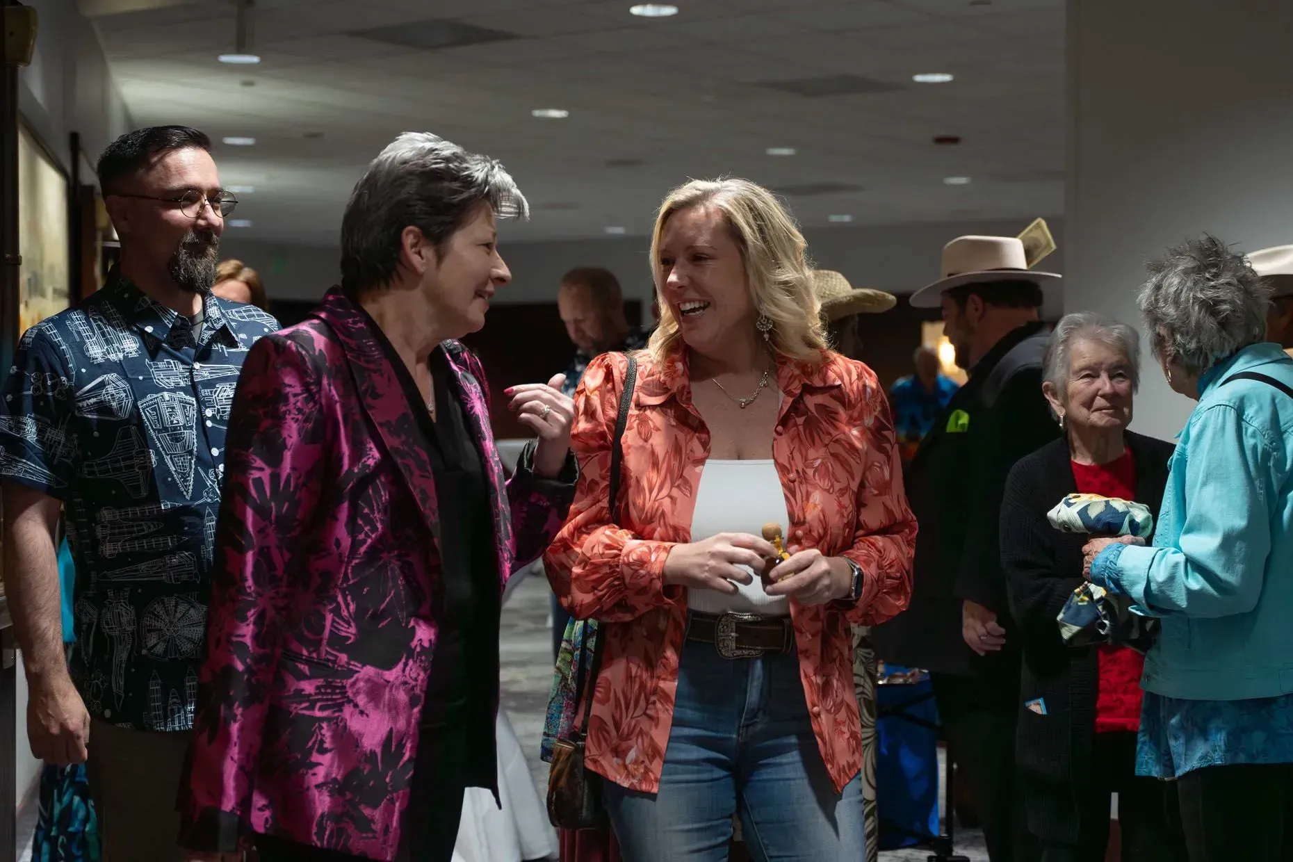 Two women chatting in a hallway, one in pink jacket, the other in an orange shirt. Others stand nearby.