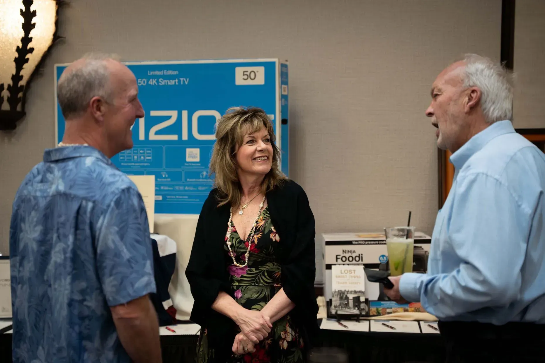 Three people conversing near a Vizio TV display. Woman in floral dress smiles; men in blue shirts gesture.