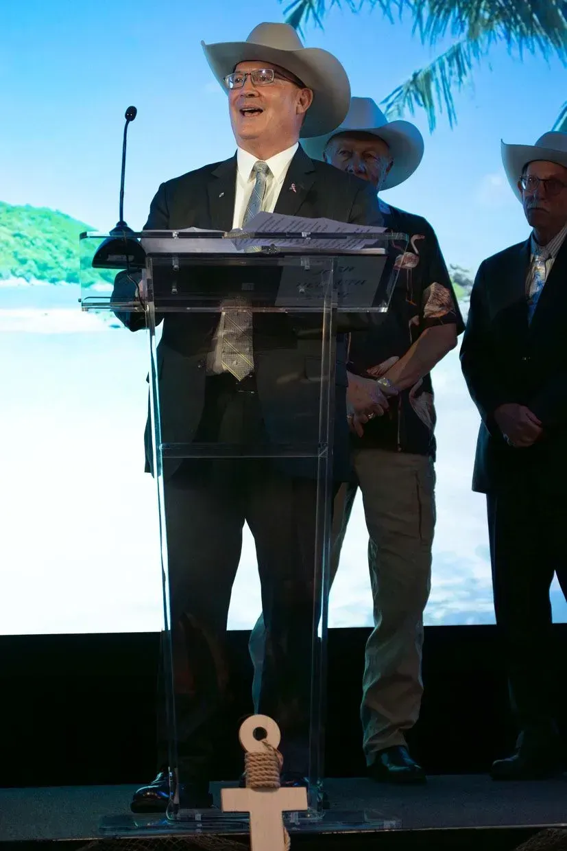 Man in cowboy hat speaks at a podium. Two other men in hats stand behind him.