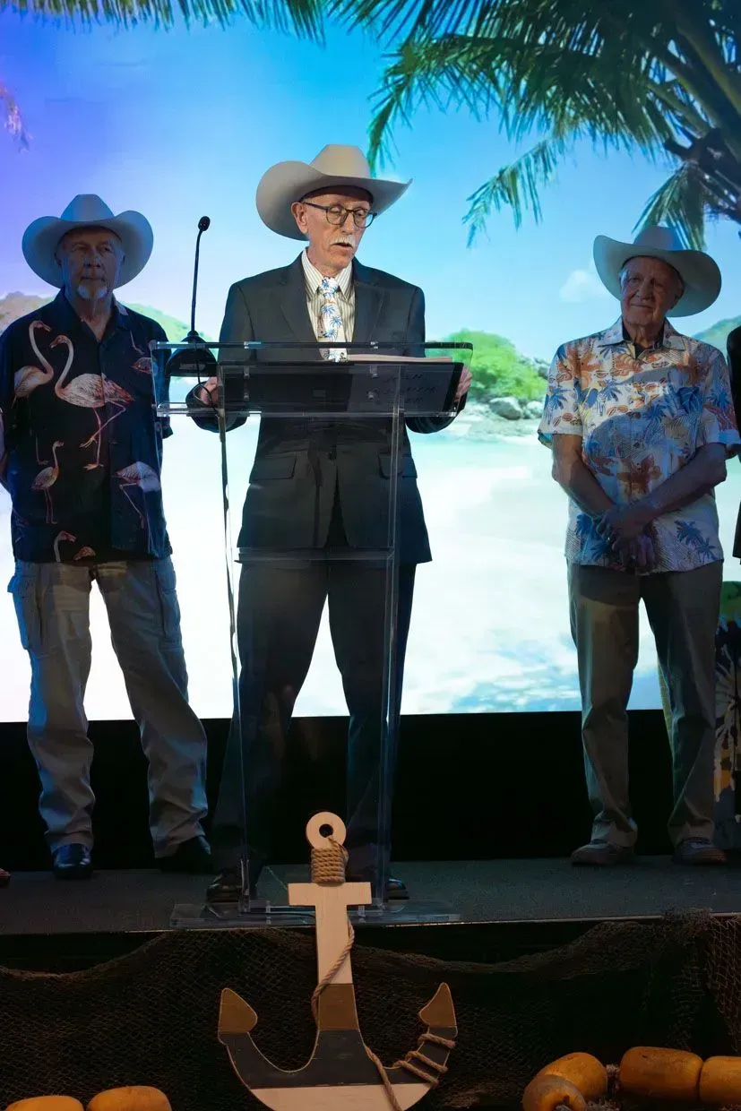 Three men in cowboy hats on a stage with a nautical backdrop, one speaking at a podium.