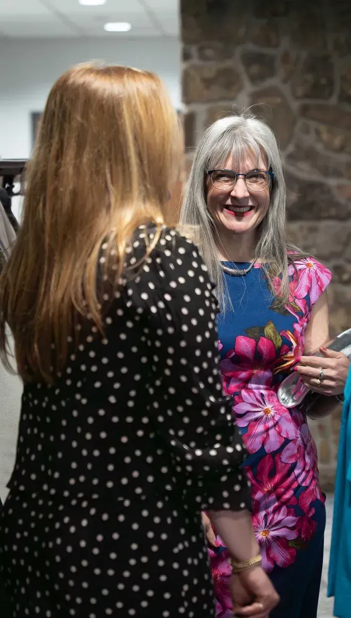 Woman in polka dots faces a smiling woman in a floral dress and glasses, inside a building.