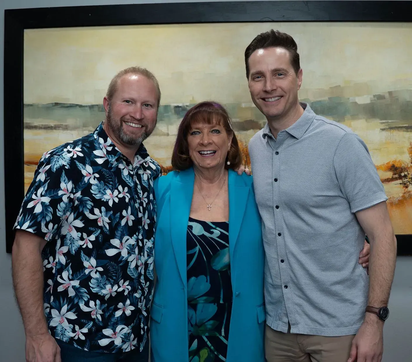 Three people posing, smiling in front of a painting. Two men, one with floral shirt. Woman in blue blazer.