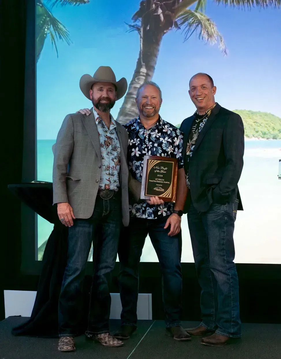 Three men in casual attire pose with an award in front of a beach backdrop.