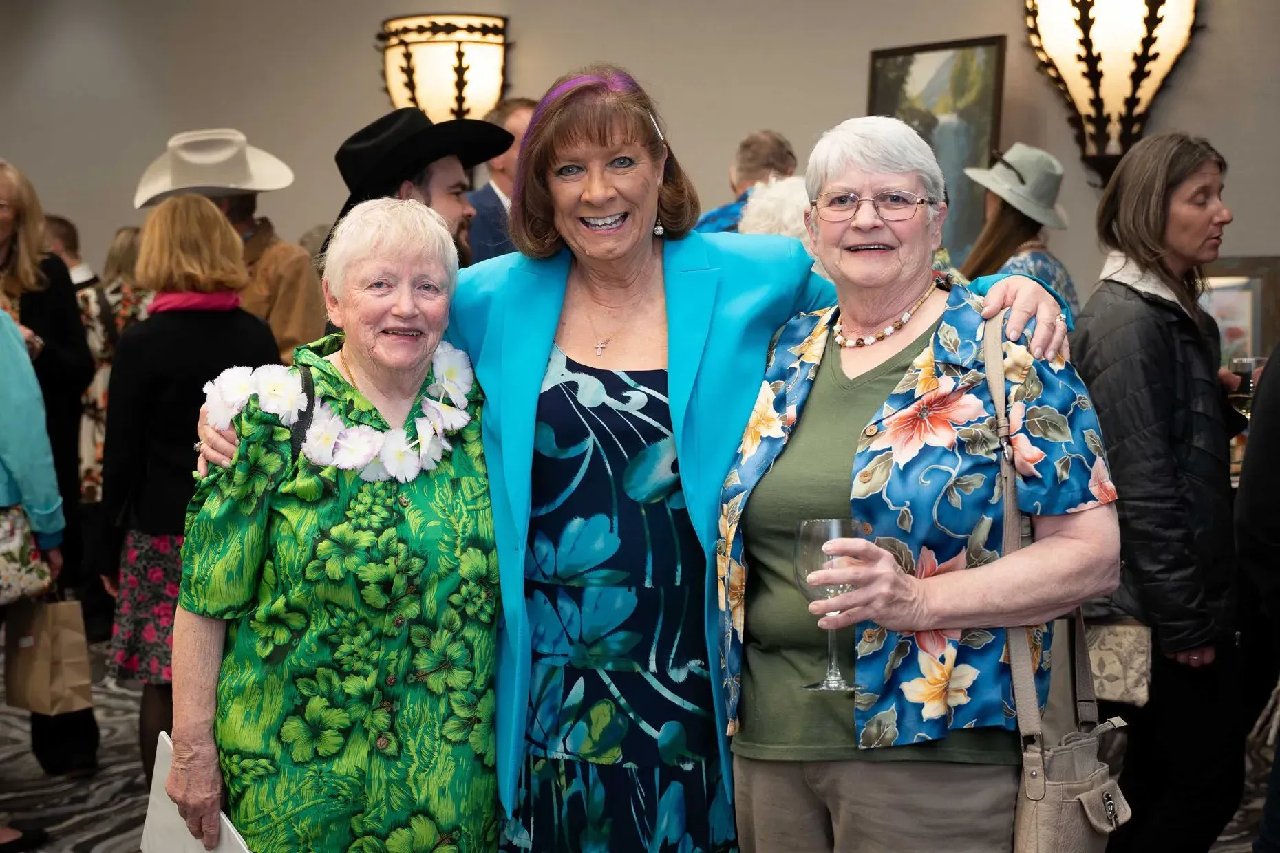 Three smiling women pose at an event. Woman in teal jacket has arms around the other two.