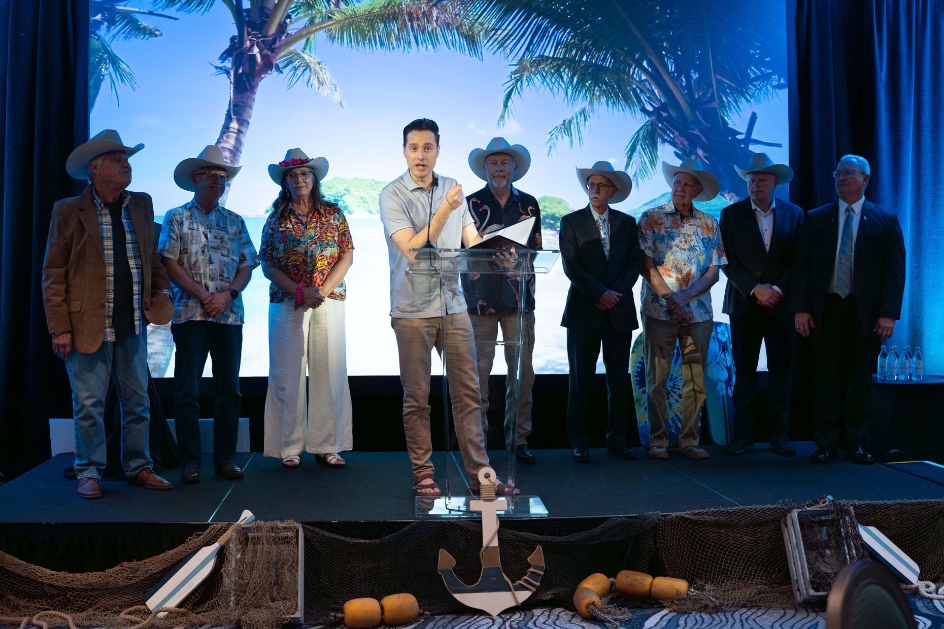 A man speaks at a podium with nine people in cowboy hats. Beach setting with blue backdrop and decor.