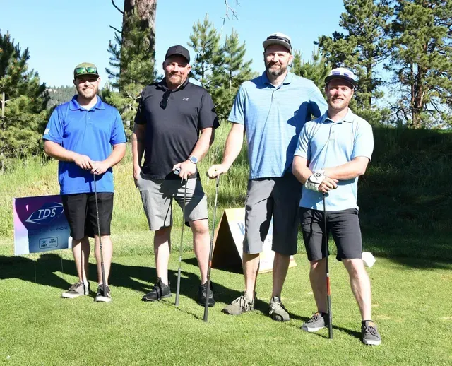 Four men posing on a golf course, smiling, holding clubs. Blue, black, and gray shirts, shorts, hats.