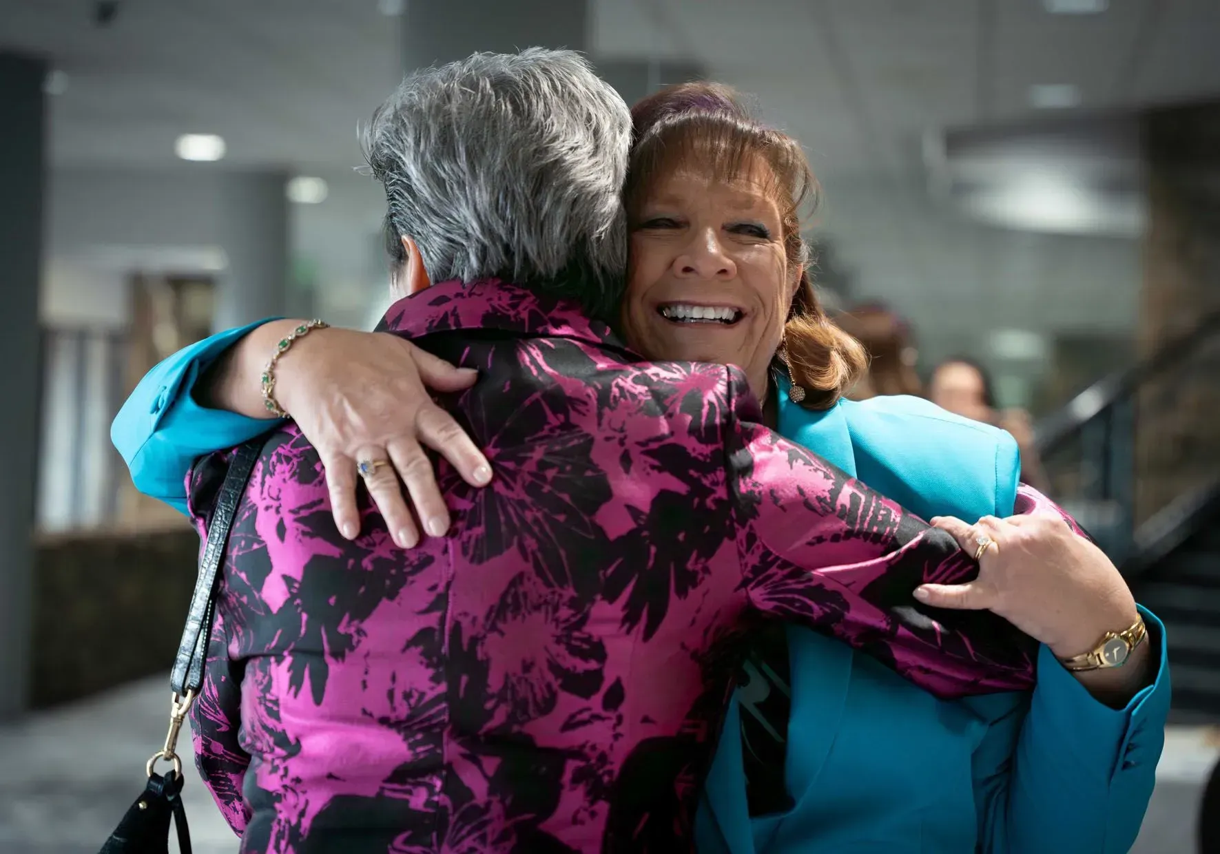 Two women in a warm embrace; one in magenta, one in blue, smiling. Interior setting.