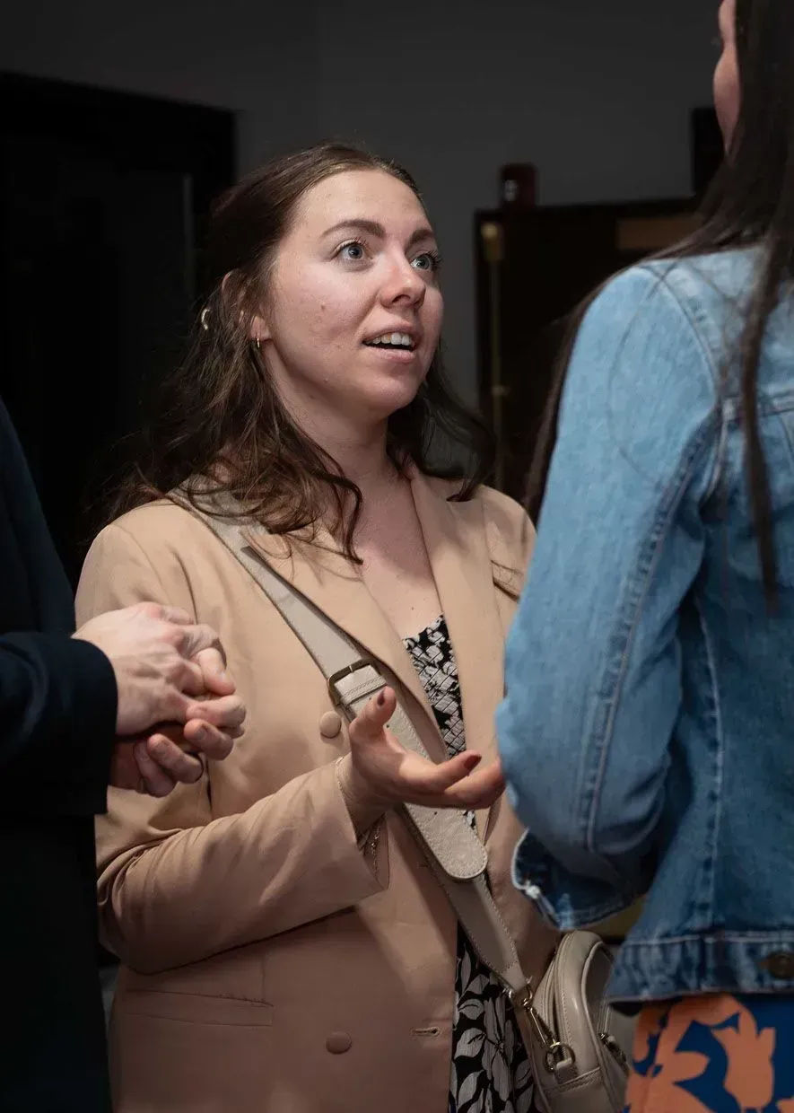 Woman in tan blazer talks to two others at a gathering.