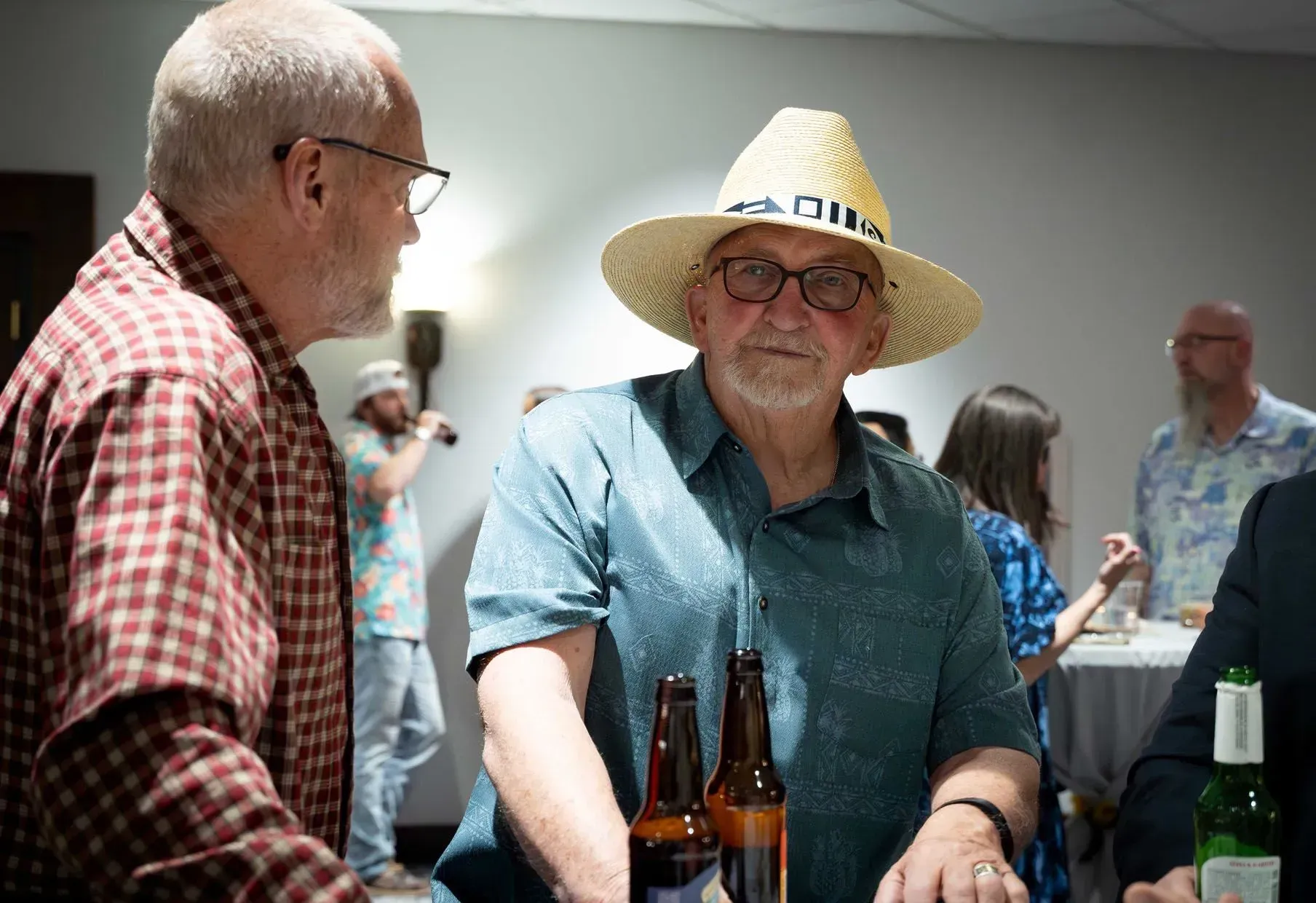 Two men in a gathering, one in a straw hat, bottles on the table, others in the background.