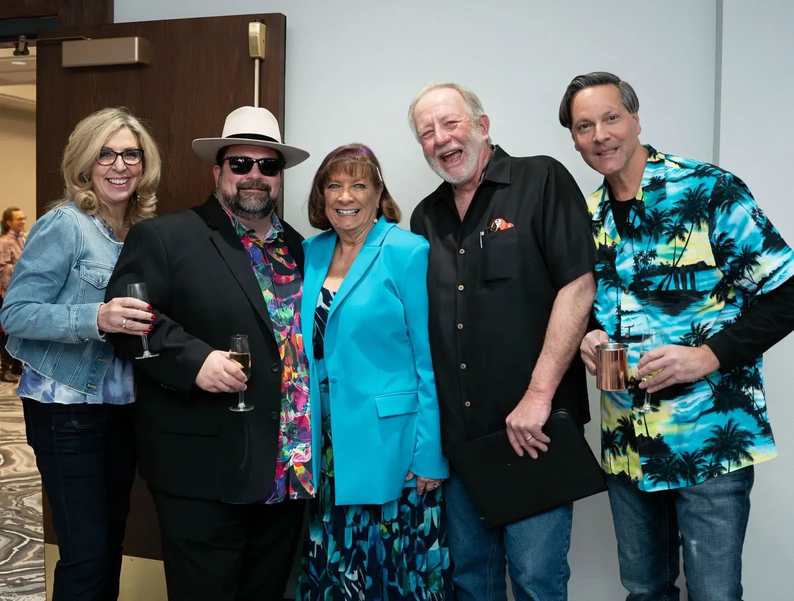 Five people smiling and posing for a photo indoors; a woman in a blue blazer, another in a Hawaiian shirt.
