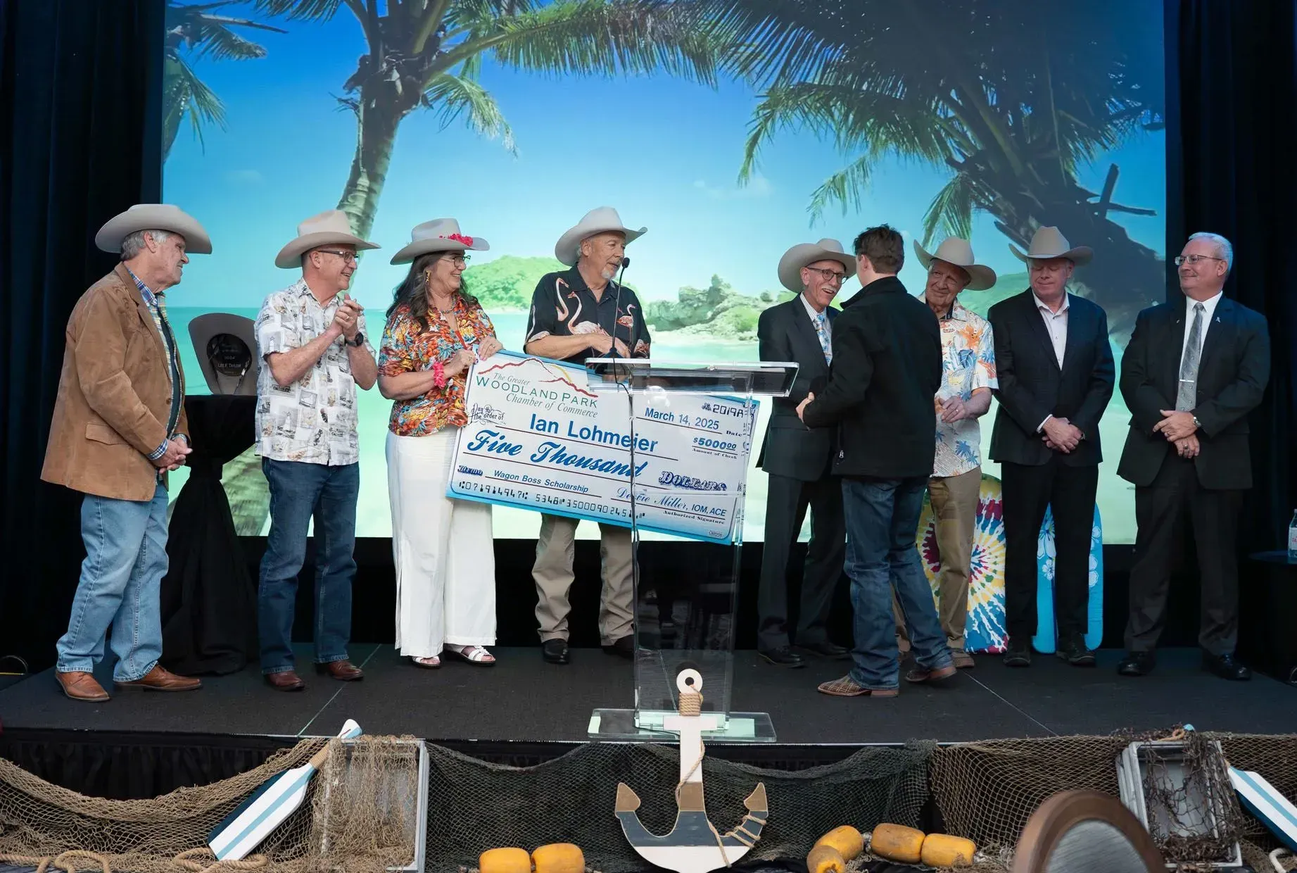 Group of people in cowboy hats on stage holding a large check with a tropical backdrop.