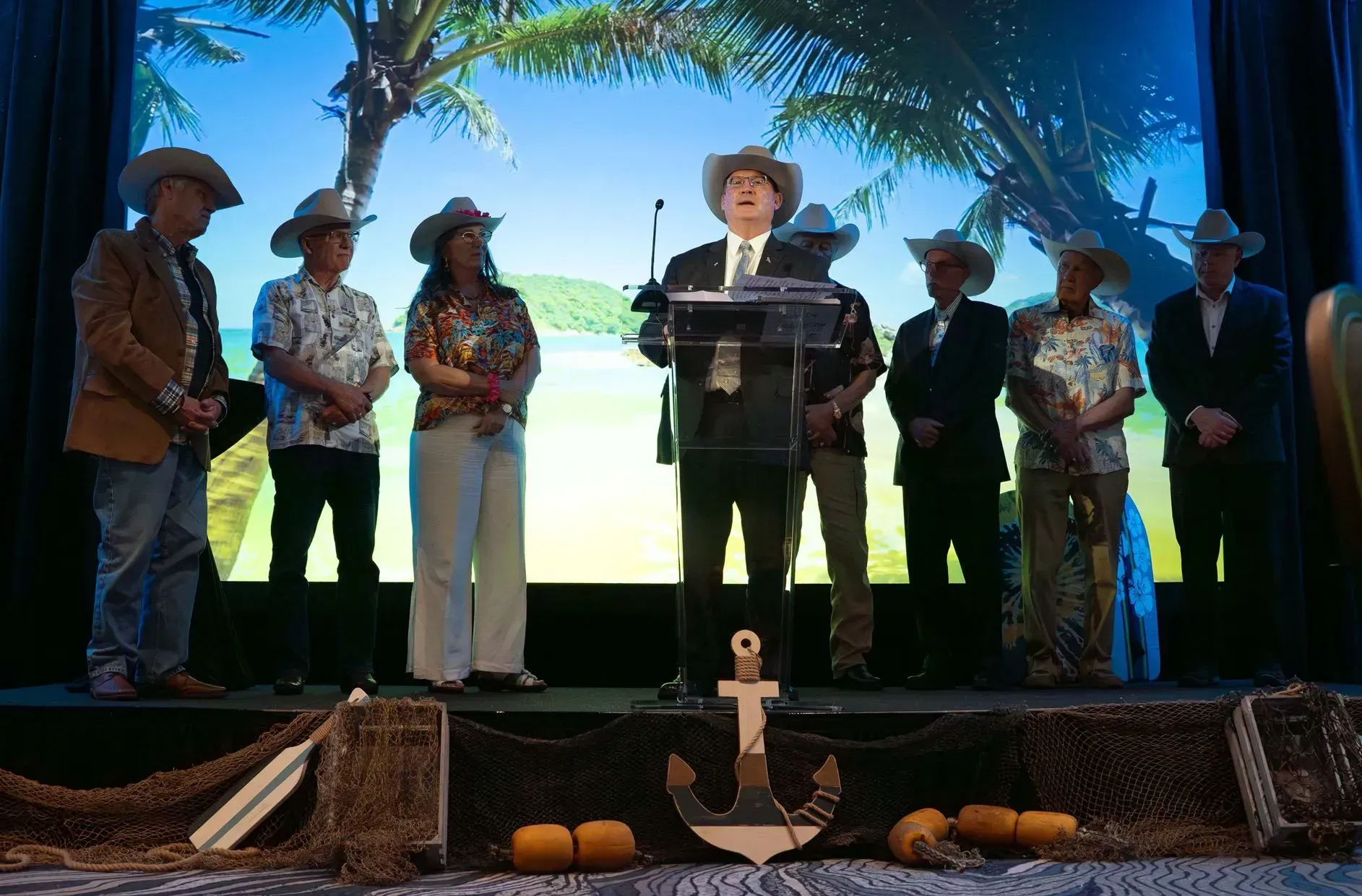 Group of people in cowboy hats on stage, one speaking at a podium, tropical backdrop.