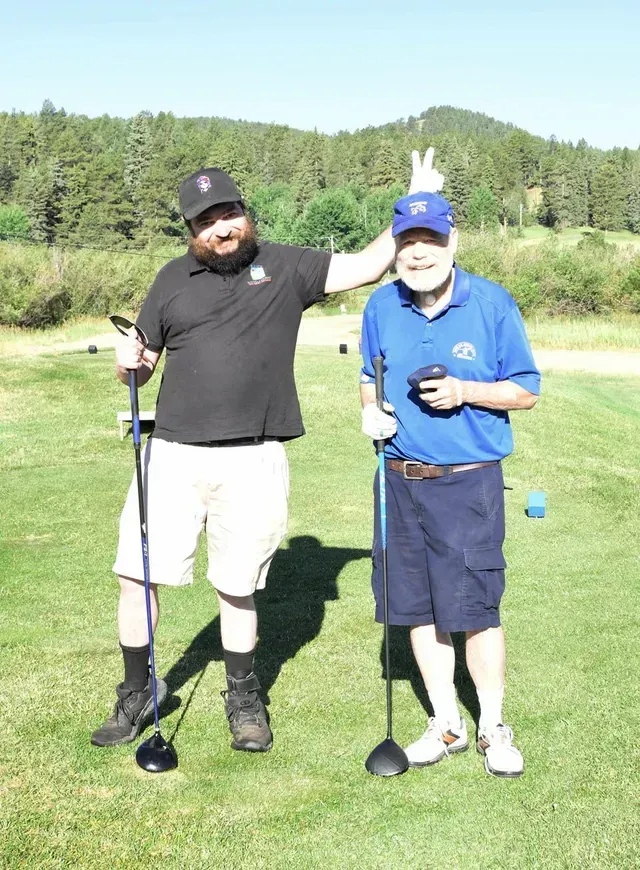 Two men on a golf course. One makes a bunny ear gesture on the other’s head. Sunny day.