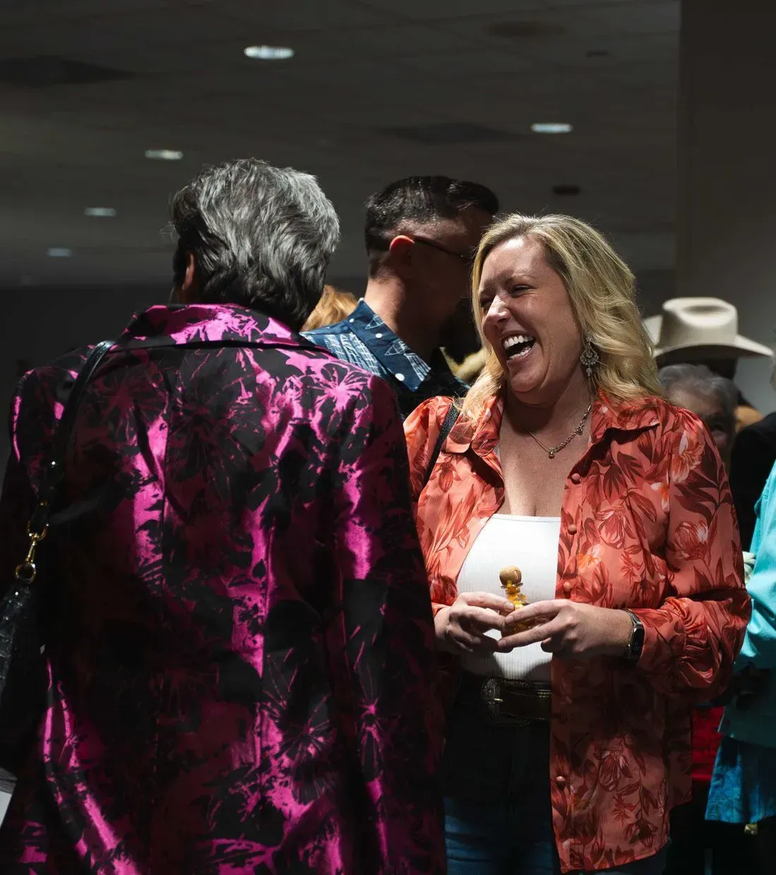 Woman in orange jacket laughs, talking with another woman in a purple jacket. Indoors.