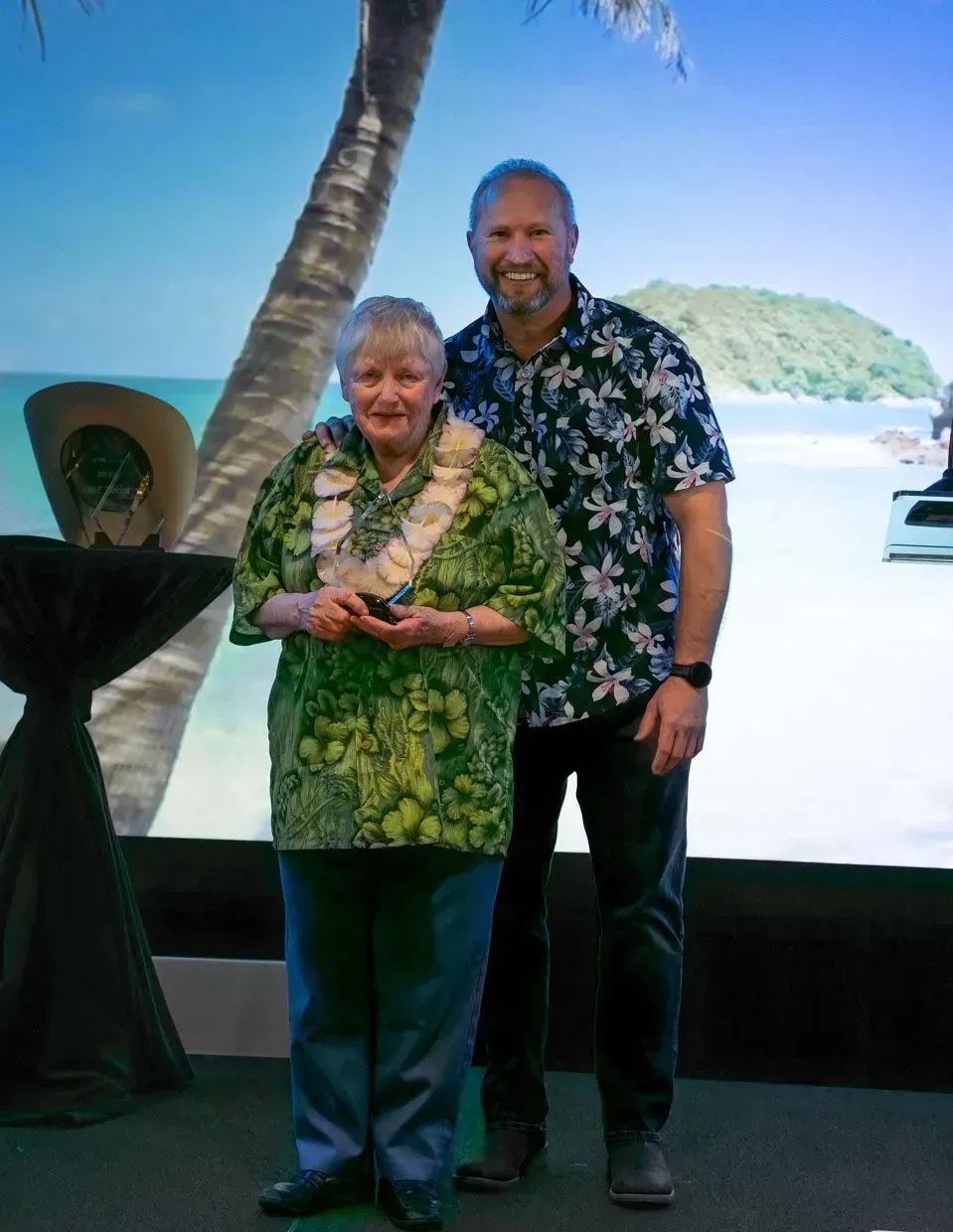 Woman in green shirt and lei, receives award, with man in floral shirt; tropical background.