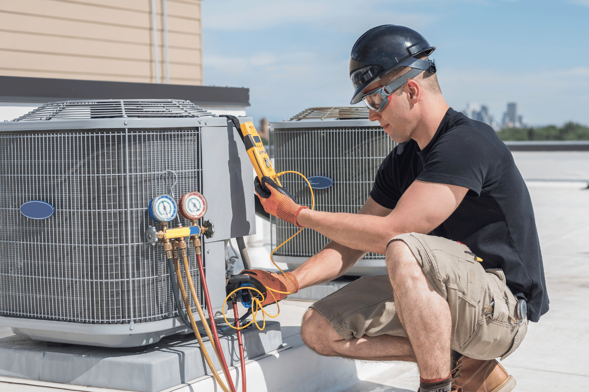 HVAC technician inspecting an AC unit on a rooftop, wearing a hard hat, safety glasses, and gloves.