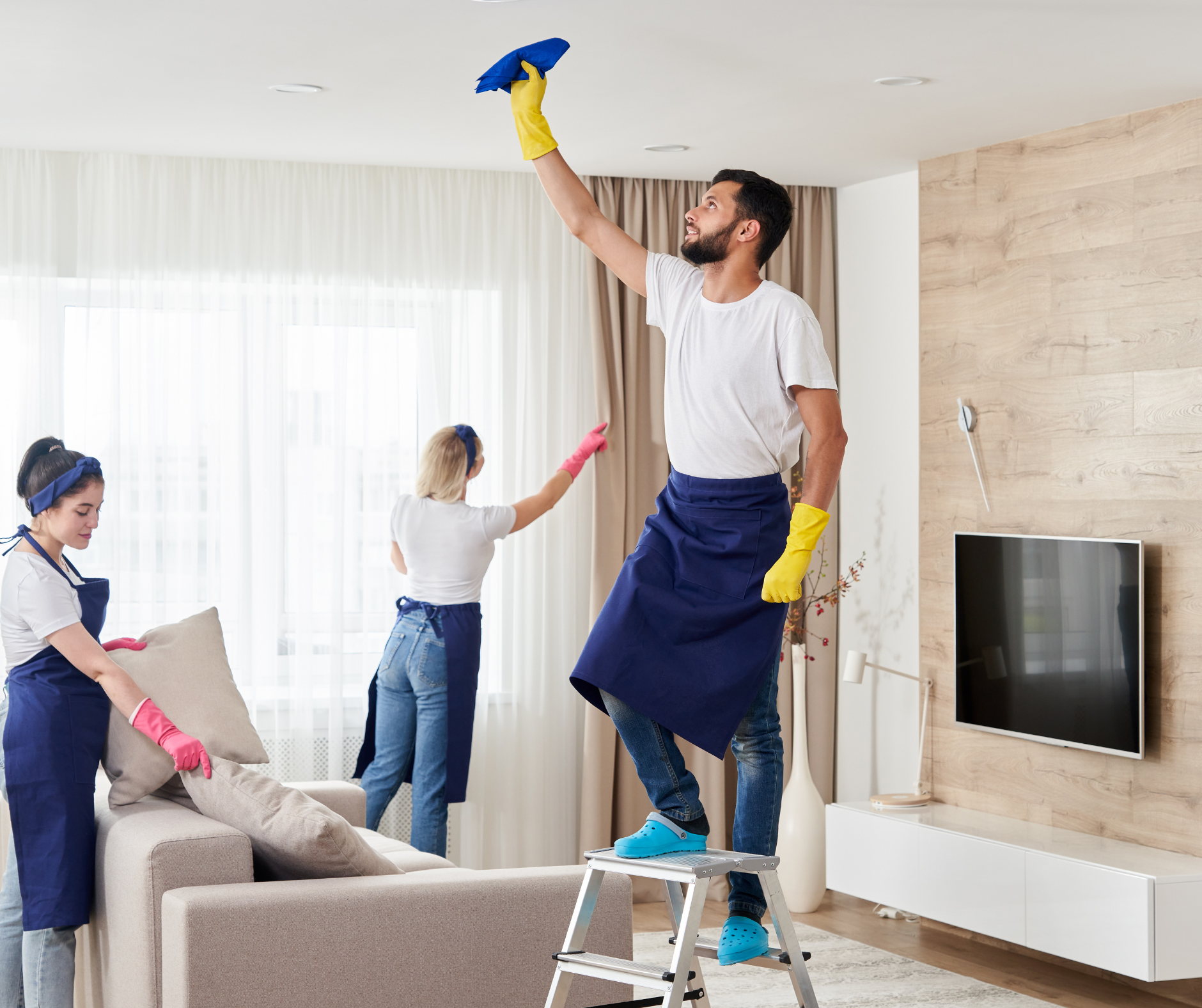 Three people cleaning a living room: one on a ladder cleaning the ceiling, two dusting furniture.