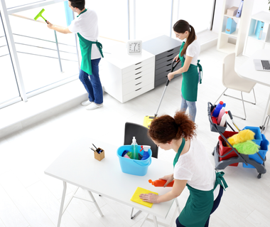 Three people cleaning an office: one window cleaning, one mopping, one wiping a table; all wearing aprons.