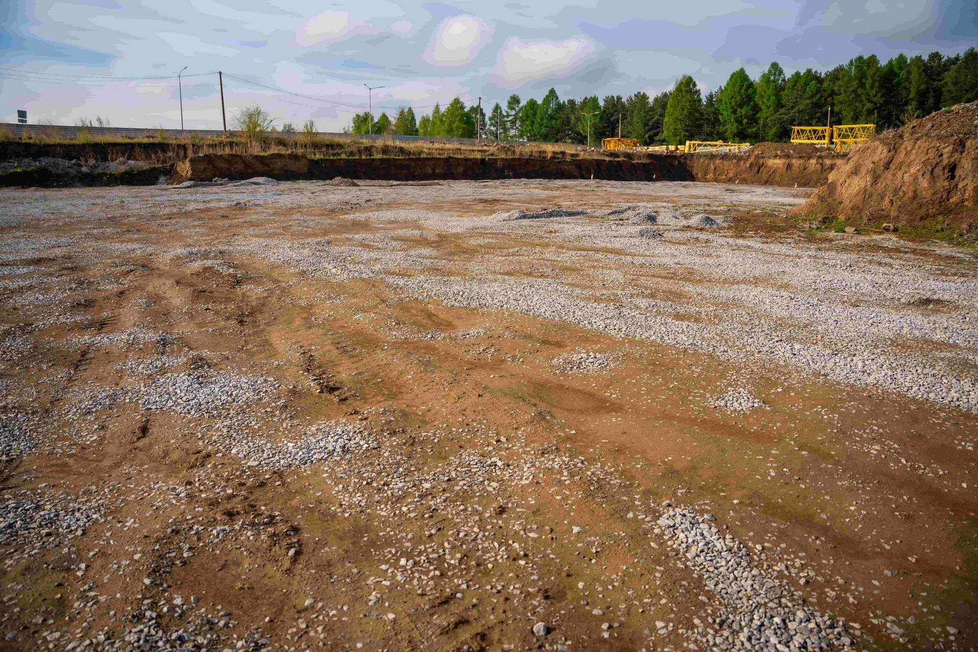 Wide muddy construction site with gravel, a line of trees, and a cloudy sky in the distance