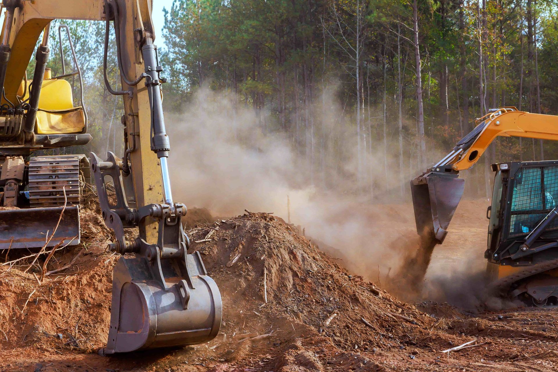 Two excavators digging and moving dirt at a dusty construction site in a wooded area