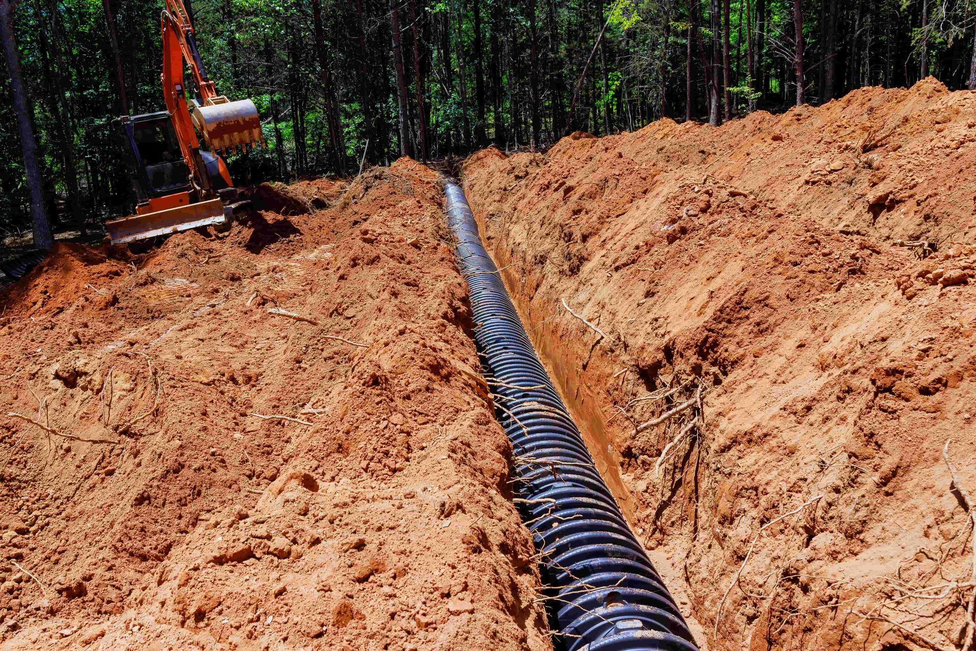 Black drainage pipe laid in a dirt trench at a wooded construction site, with a worker in the background