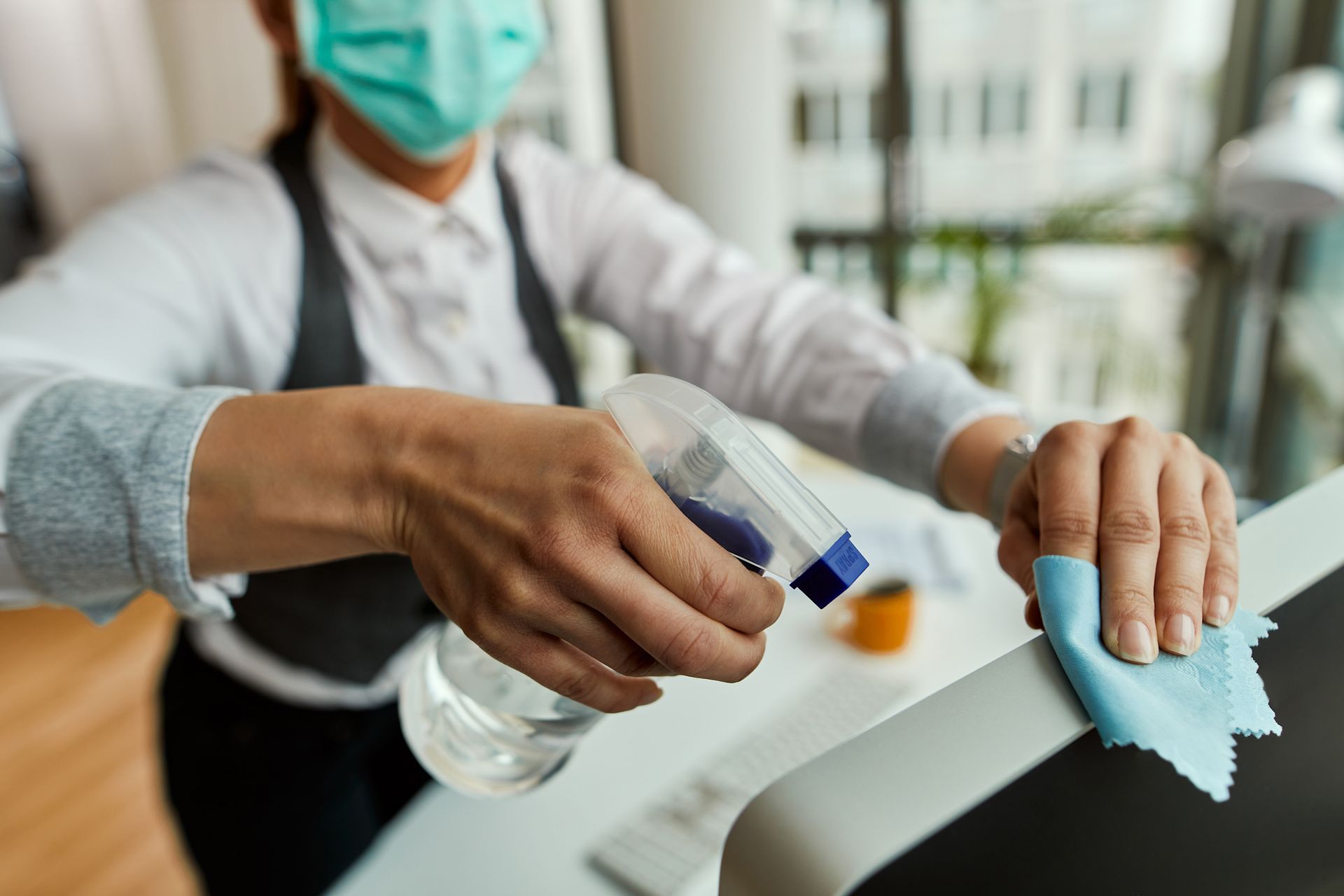 Person in mask disinfecting a surface with spray and cloth in a bright office