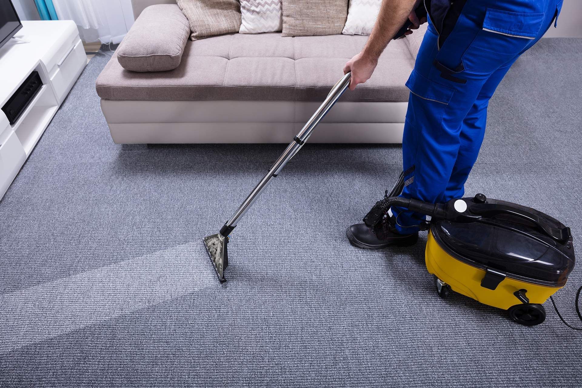 Person in blue overalls vacuuming gray carpet in a living room; yellow vacuum cleaner visible.