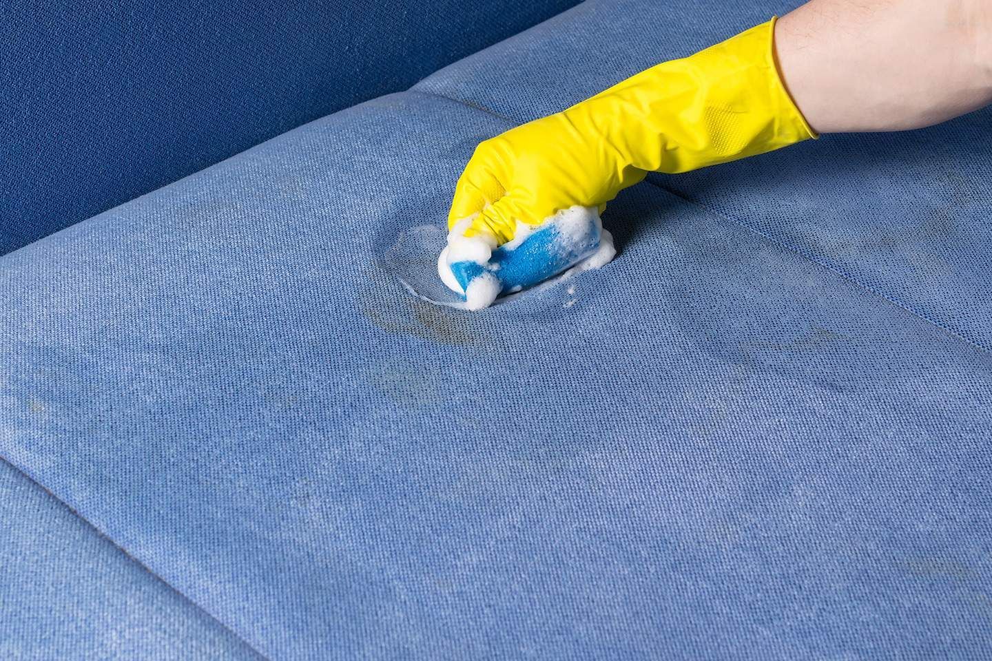 Person wearing yellow gloves cleaning a blue couch with a sponge.