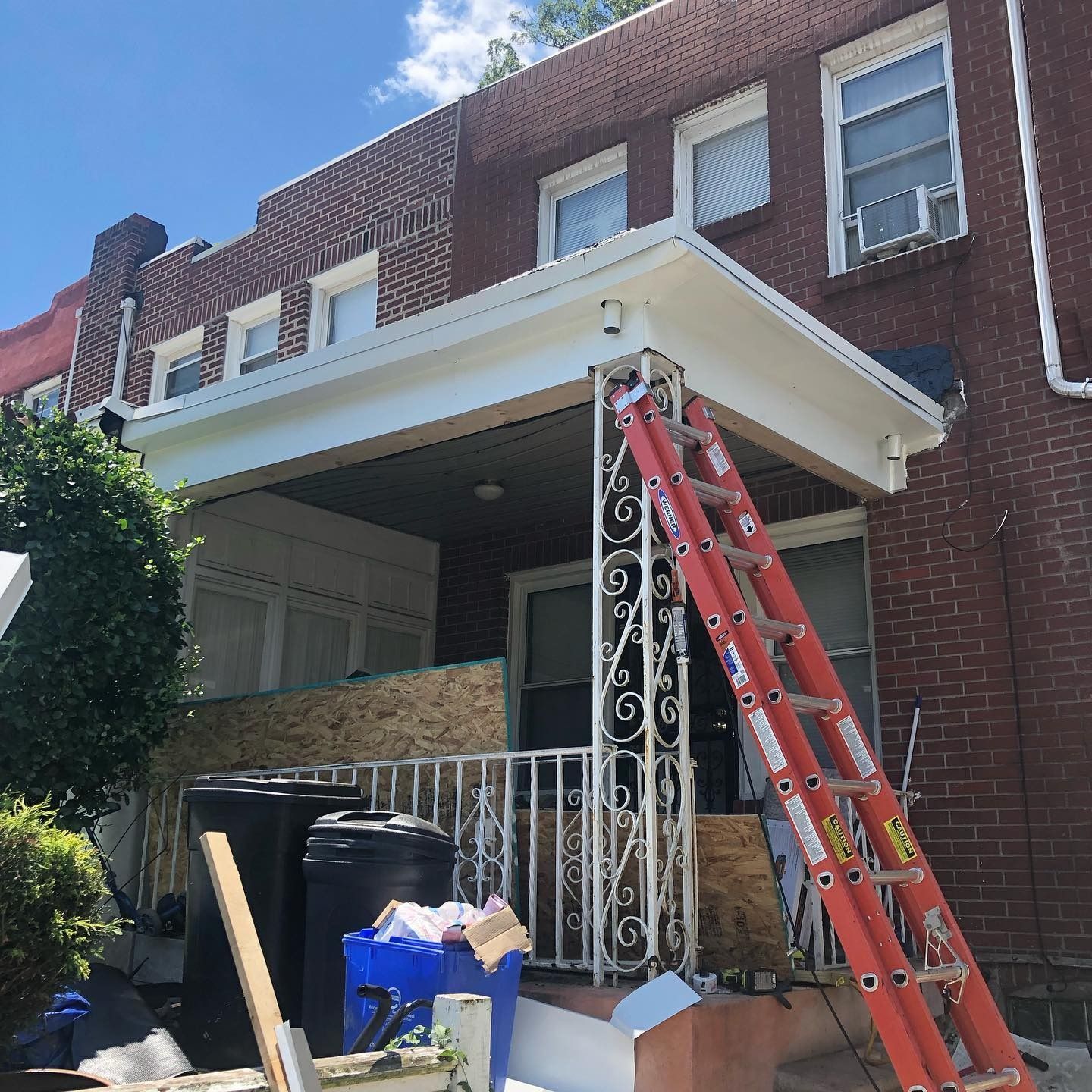 Red ladder leaning against a brick house porch with trash bins and renovation debris below.