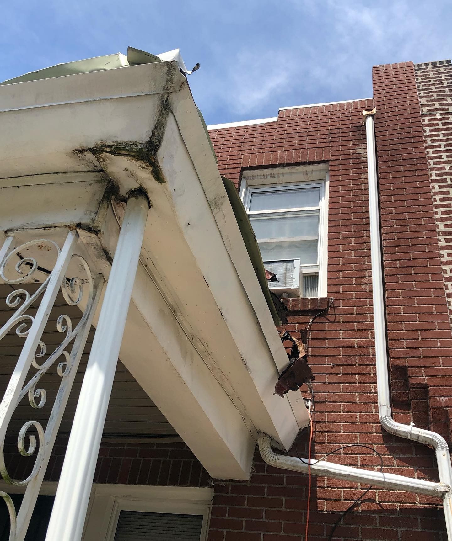 Damaged awning beside a brick building, with a window and downspout above a porch railing.
