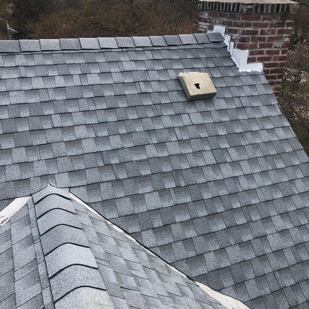 Gray shingled roof with a small vent and brick chimney against a rooftop backdrop
