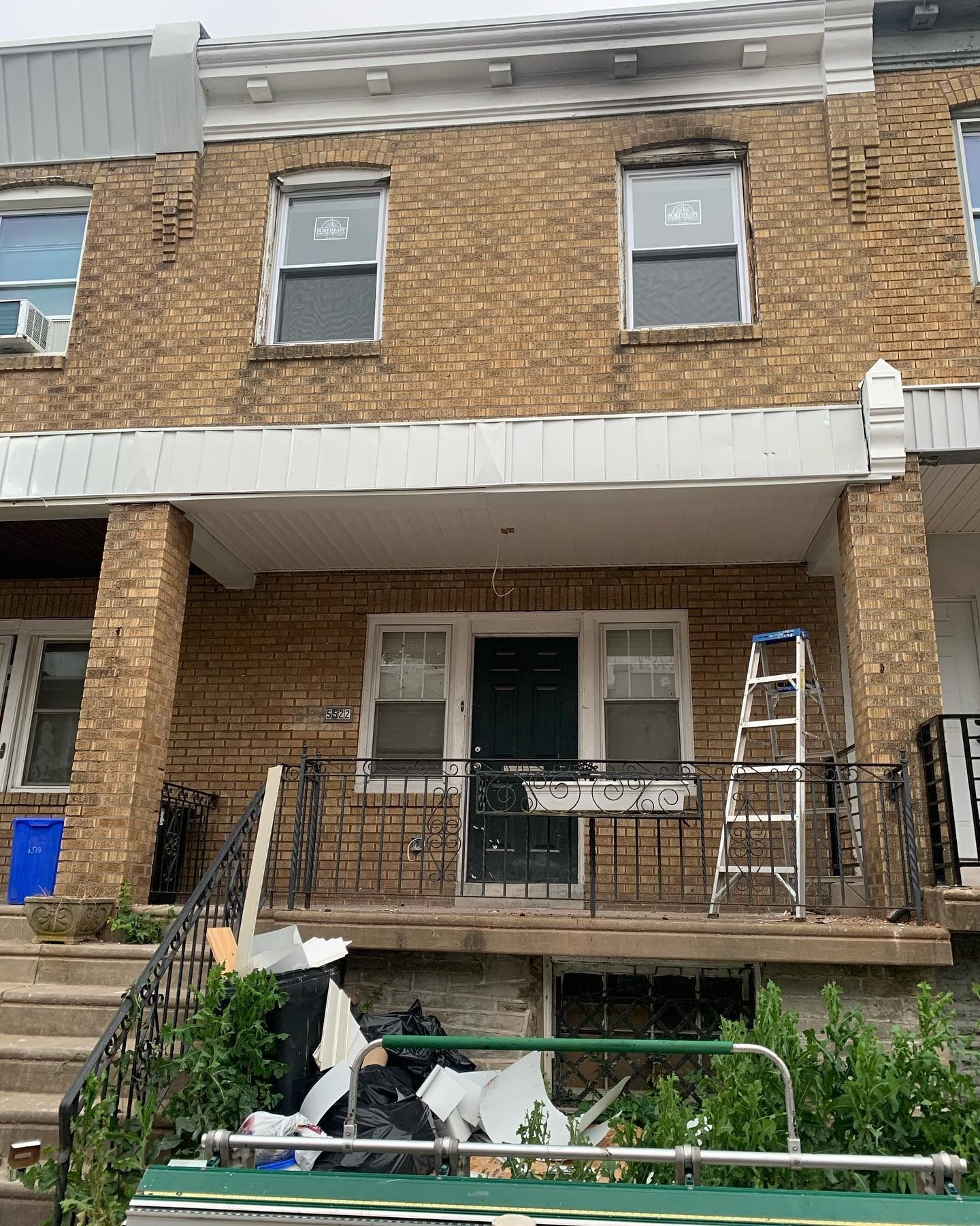 Brick row house with front porch, railing, ladder, and basement opening under renovation